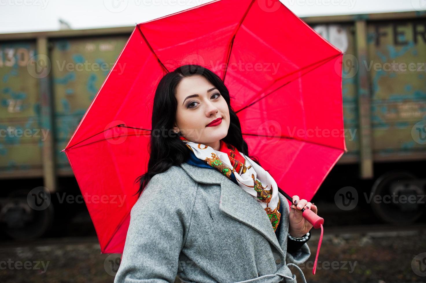 ragazza bruna in cappotto grigio con ombrello rosso nella stazione ferroviaria. foto