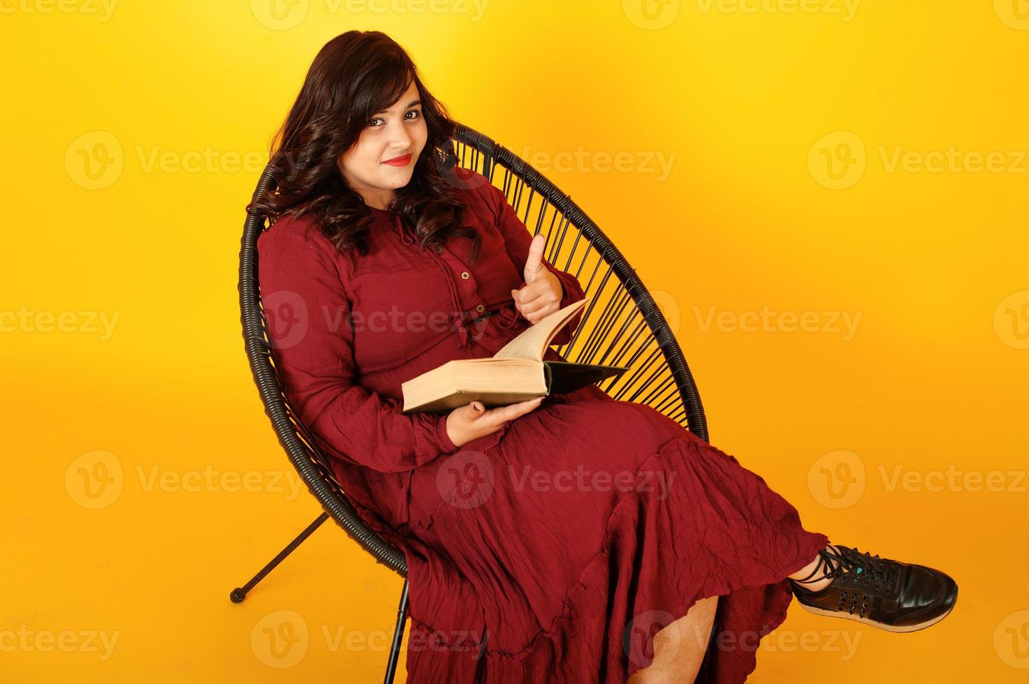 attraente donna del sud asiatico in abito rosso intenso poste in studio su sfondo giallo seduto su una sedia con il libro. foto