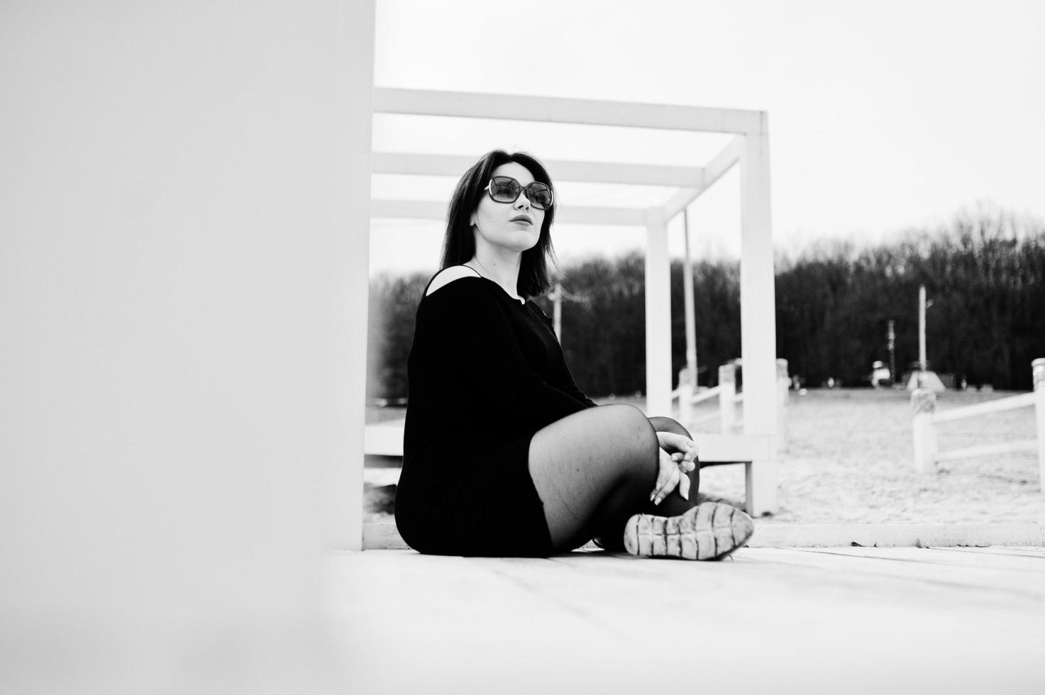 ritratto di ragazza bruna in abito nero seduto a costruzione in legno bianco. foto
