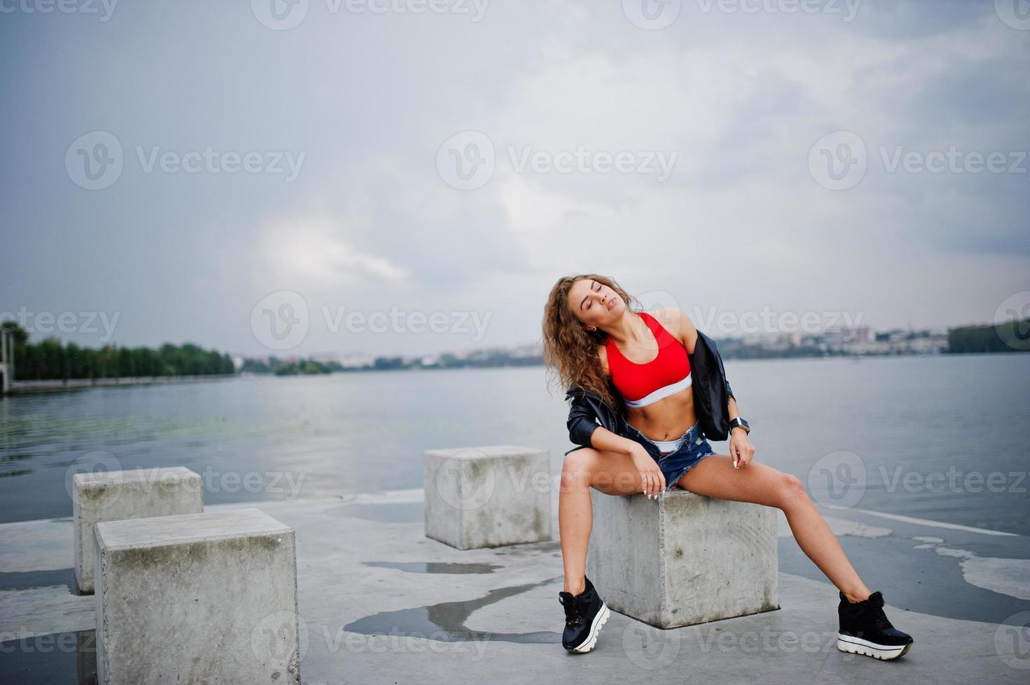 ragazza modello riccia sexy in top rosso, pantaloncini di jeans, giacca di pelle e scarpe da ginnastica poste a cubi di pietra. foto