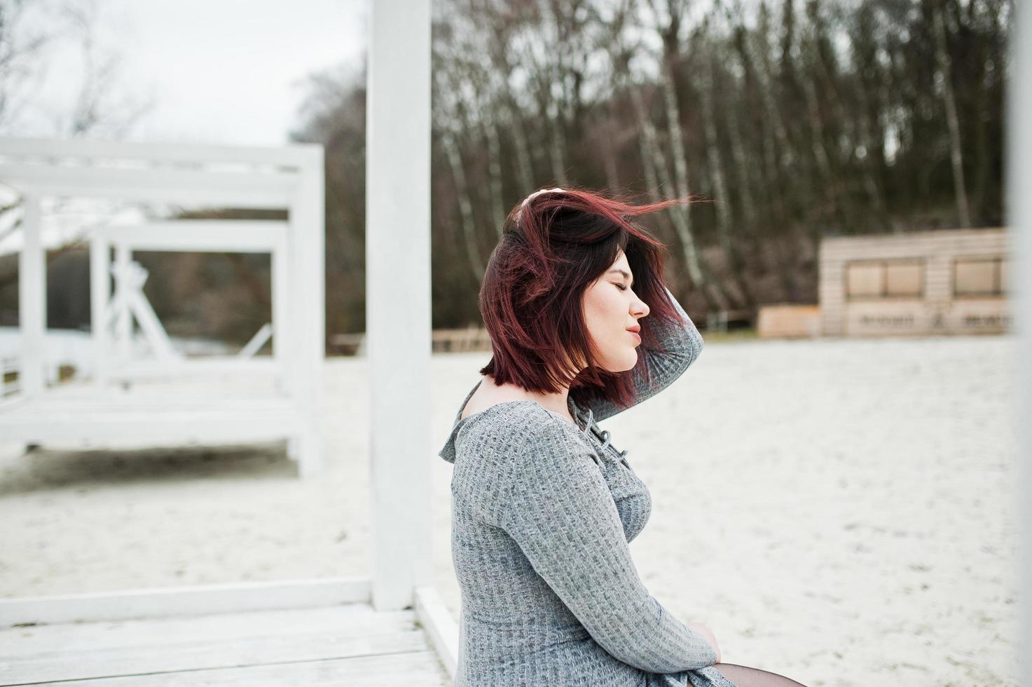 ritratto di ragazza bruna in abito grigio seduto a una costruzione in legno bianco. foto