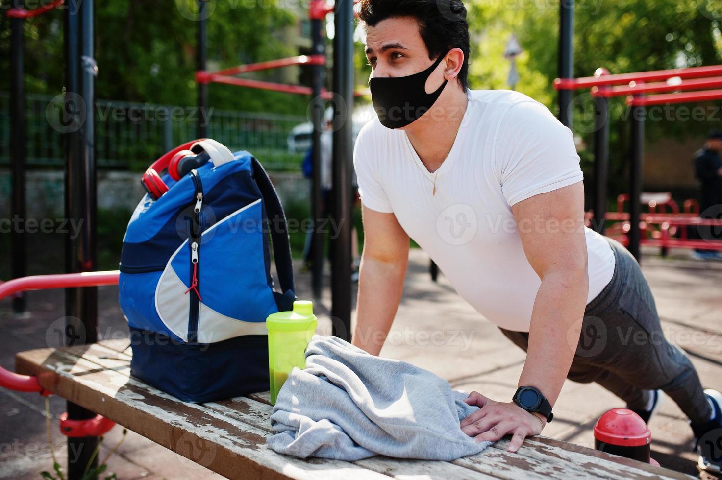 ritratto sport uomo arabo in maschera medica nera facendo esercizi di allenamento in palestra all'aperto durante la quarantena del coronavirus. foto
