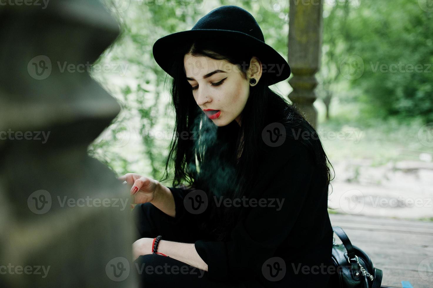 sensuale fumatrice tutta in nero, labbra rosse e cappello. donna drammatica goth che fuma sigaretta sottile. foto