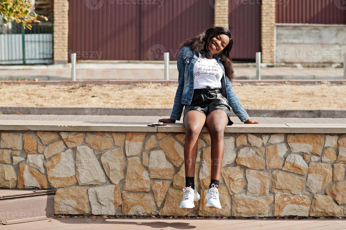 modello slim afroamericano dalla pelle scura posato in pantaloncini neri e giacca di jeans con telefono cellulare. foto