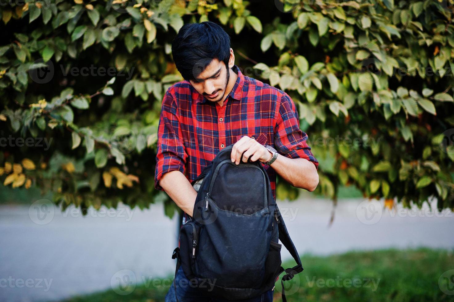 giovane studente indiano in camicia a scacchi rossa e jeans con zaino in posa in strada. foto