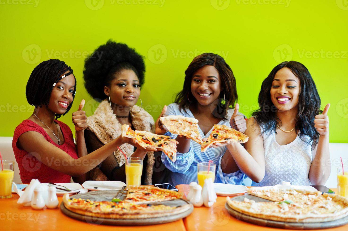 quattro giovani ragazze africane in un ristorante dai colori vivaci che tengono in mano le fette di pizza e mostrano il pollice in su. foto