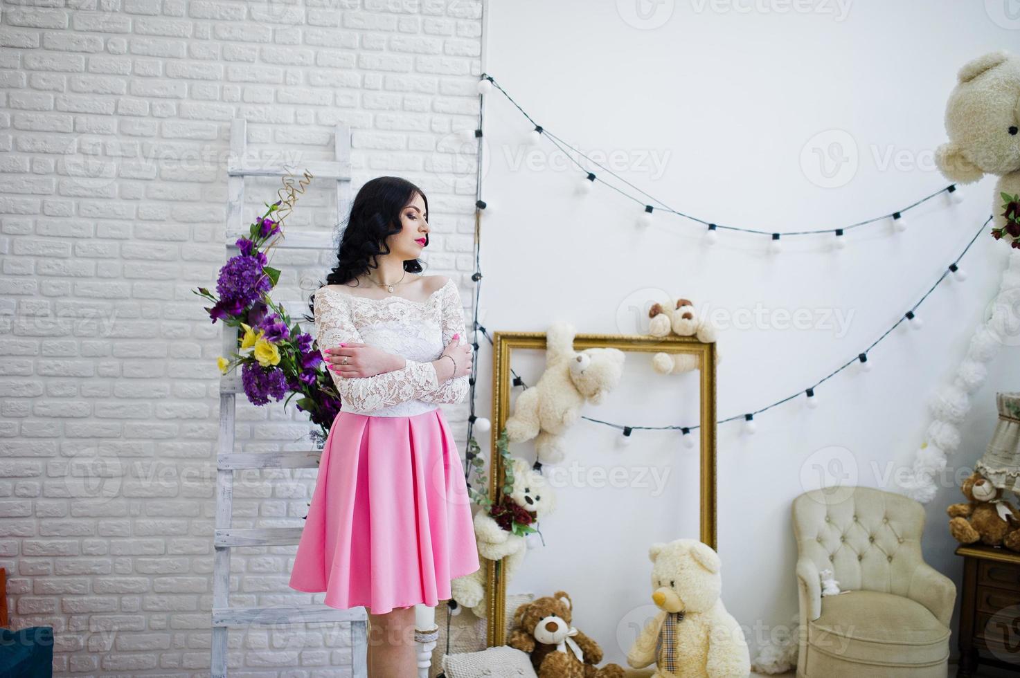 giovane ragazza bruna in gonna rosa e camicetta bianca poste al coperto contro la stanza con l'orso dei giocattoli. foto