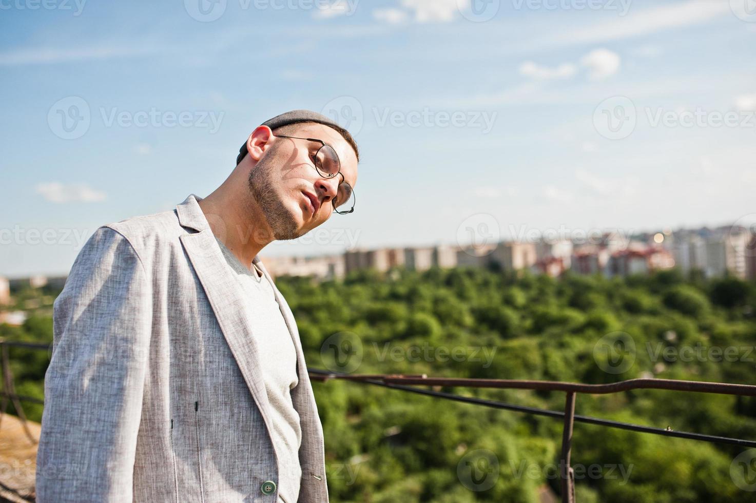 sognatore elegante uomo macho in abito grigio, cappello e occhiali in posa sul tetto. foto