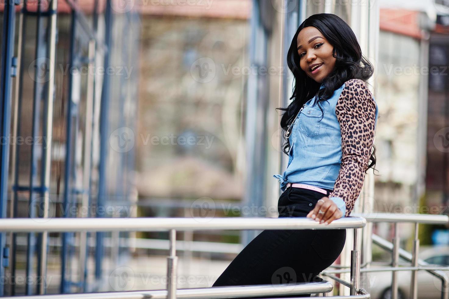 hipster ragazza afroamericana che indossa una camicia di jeans con maniche di leopardo in posa in strada contro un moderno edificio per uffici con finestre blu. foto