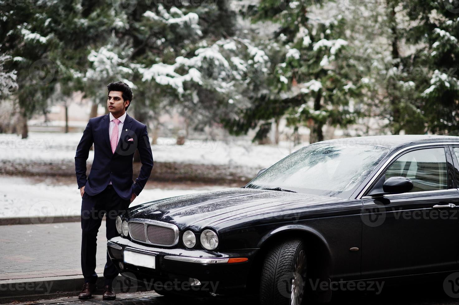 elegante modello uomo macho indiano su abito e cravatta rosa in posa contro un'auto classica nera. foto