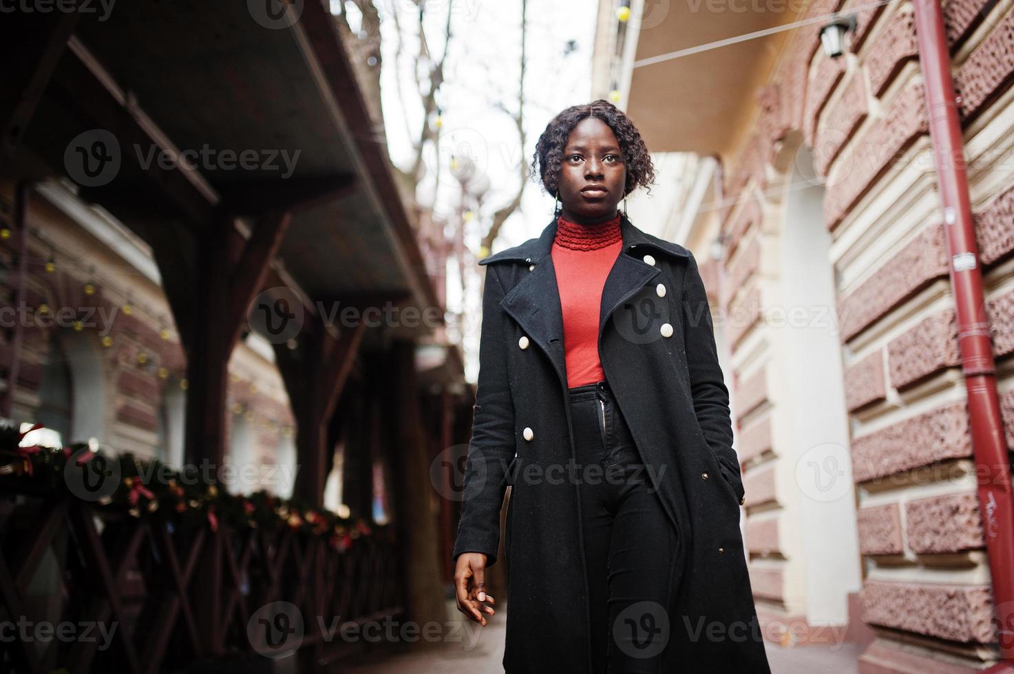 ritratto di una donna africana dai capelli ricci che indossa un cappotto nero alla moda e un dolcevita rosso che cammina all'aperto. foto