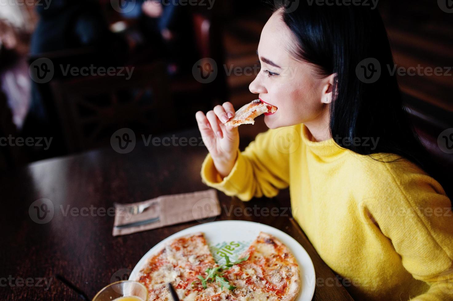 divertente ragazza bruna in maglione giallo che mangia pizza al ristorante. foto