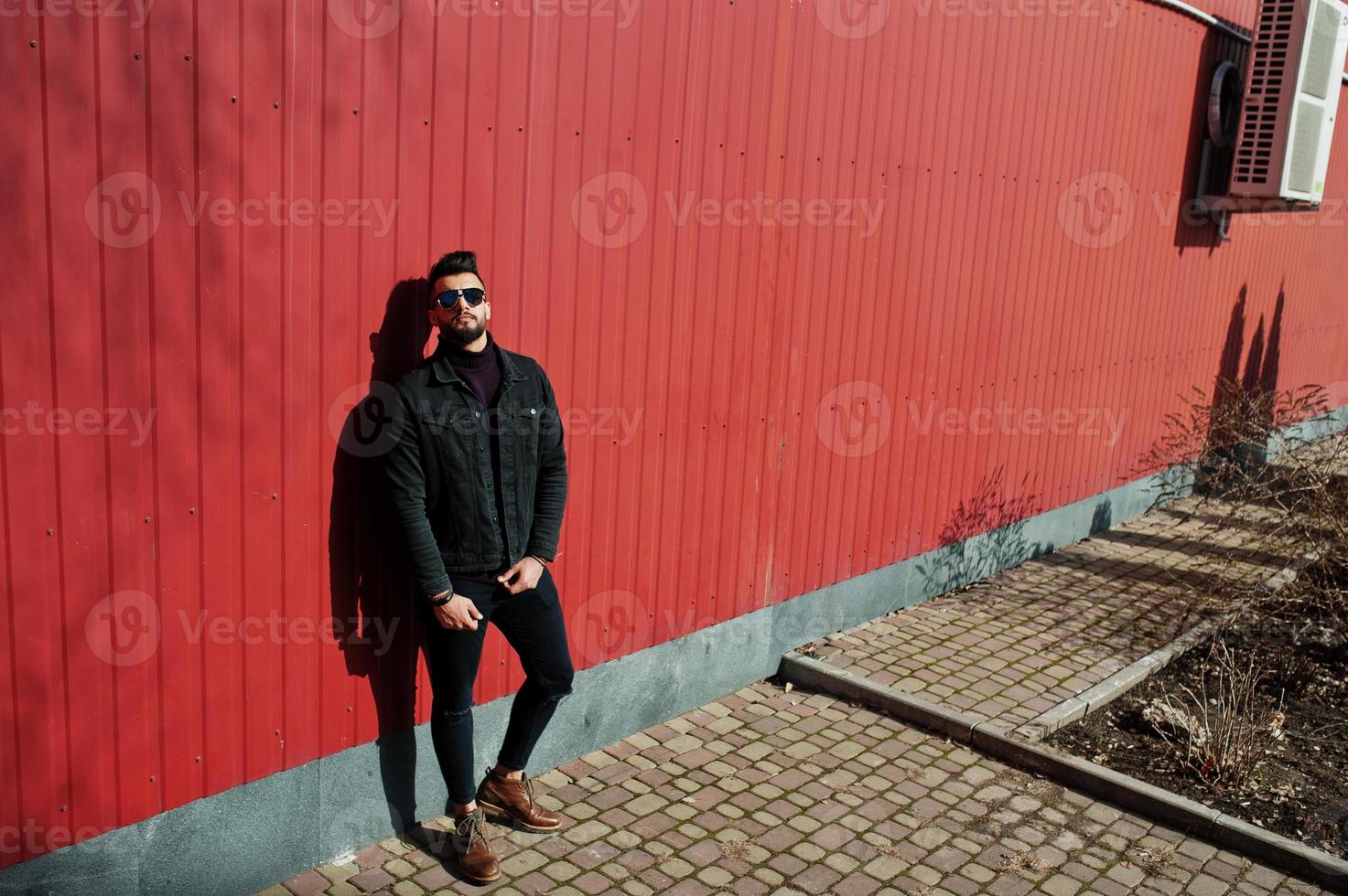 l'uomo arabo di moda indossa una giacca di jeans nera e occhiali da sole poste sullo sfondo della parete in acciaio rosso. ragazzo modello arabo elegante e alla moda. foto