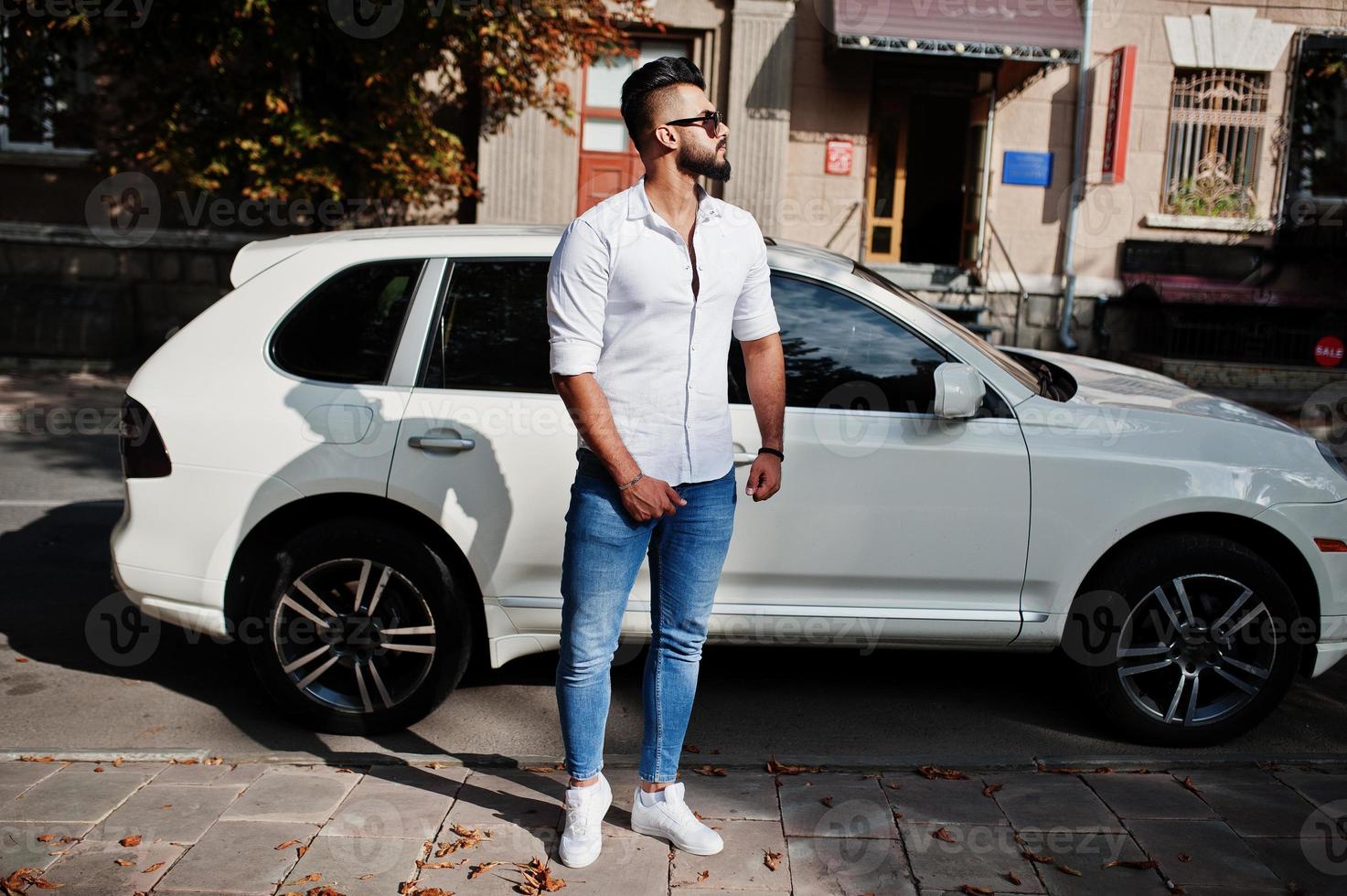 elegante uomo arabo alto modello in camicia bianca, jeans e occhiali da sole posato in una strada della città. ragazzo arabo ricco e attraente della barba contro l'automobile bianca del suv. foto