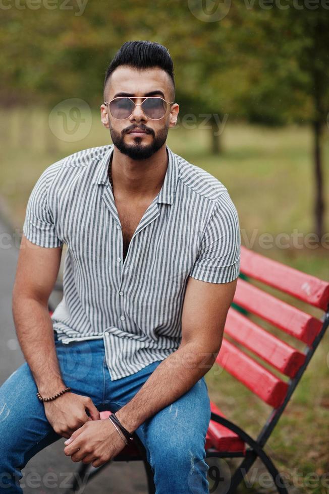 l'uomo alla moda con barba araba alta indossa camicia, jeans e occhiali da sole posati sul parco in panchina. foto