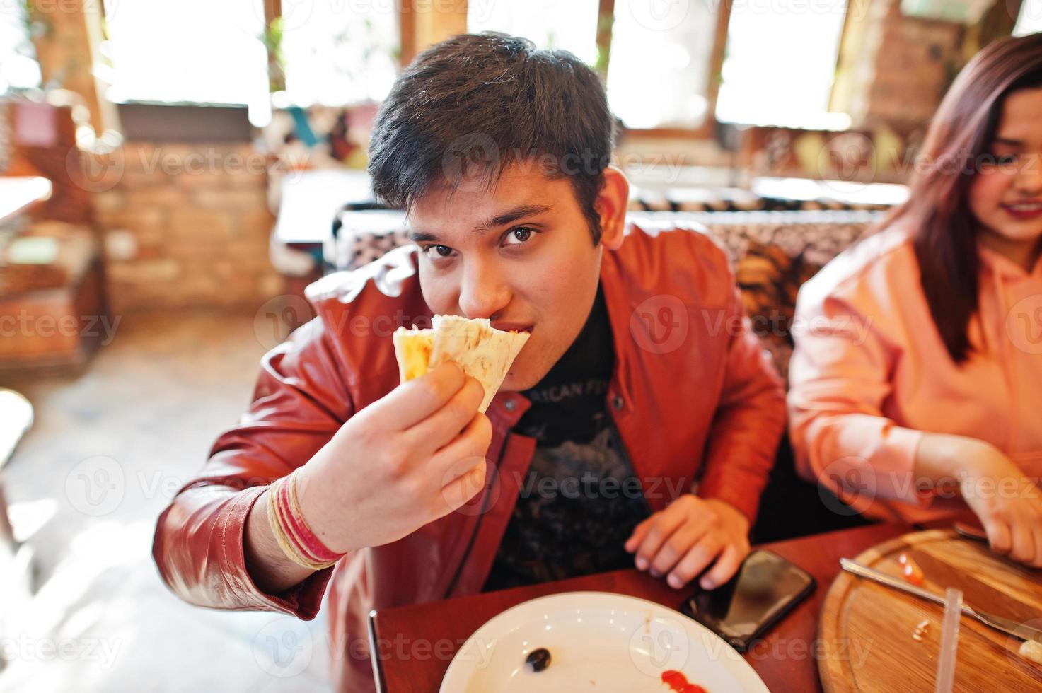 coppia di amici asiatici che mangiano pizza durante la festa in pizzeria. felici indiani che si divertono insieme, mangiano cibo italiano e si siedono sul divano. foto