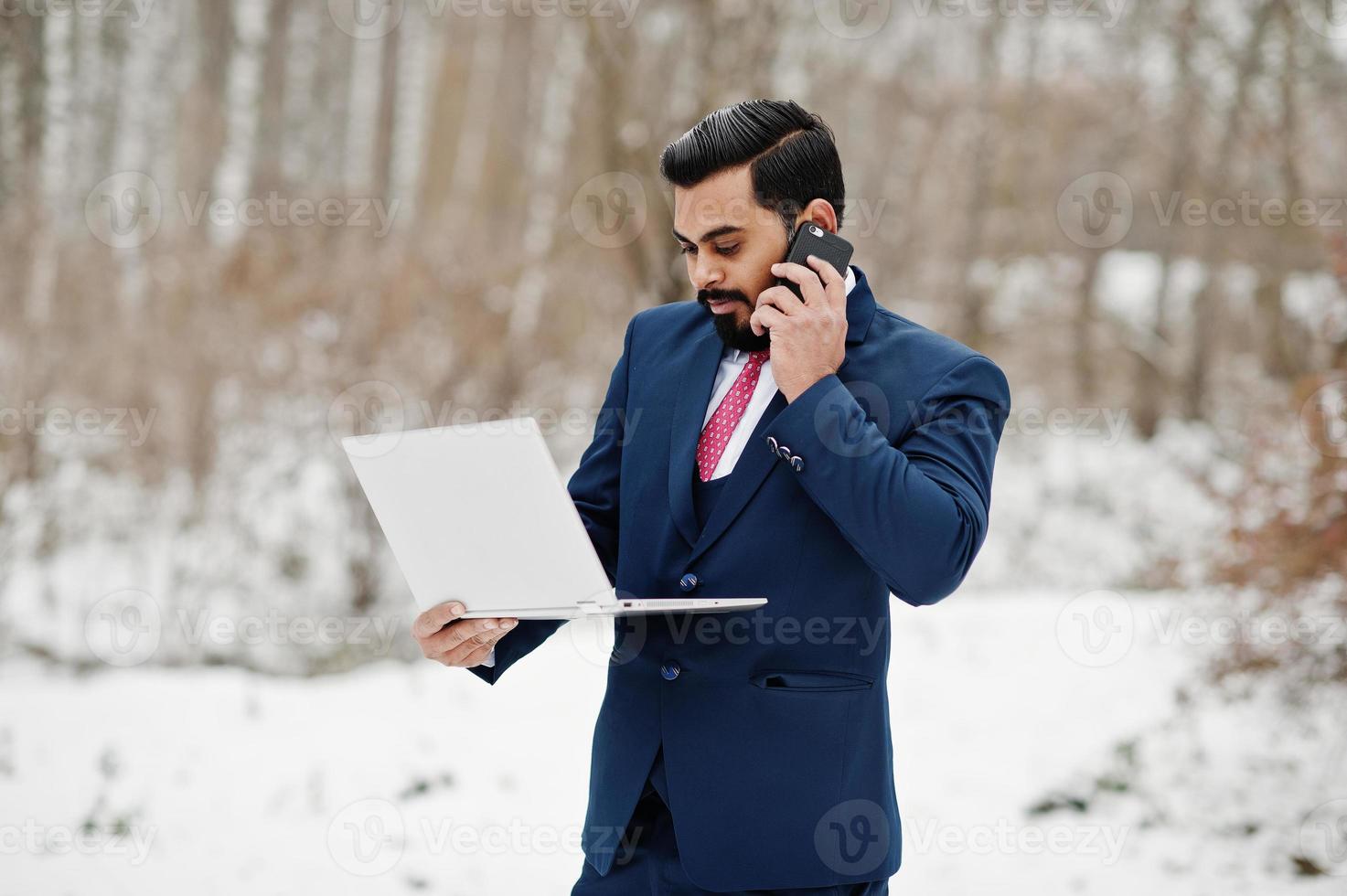 elegante uomo d'affari con barba indiana in giacca e cravatta posato in una giornata invernale all'aperto con un laptop a portata di mano, parlando sul telefono cellulare. foto