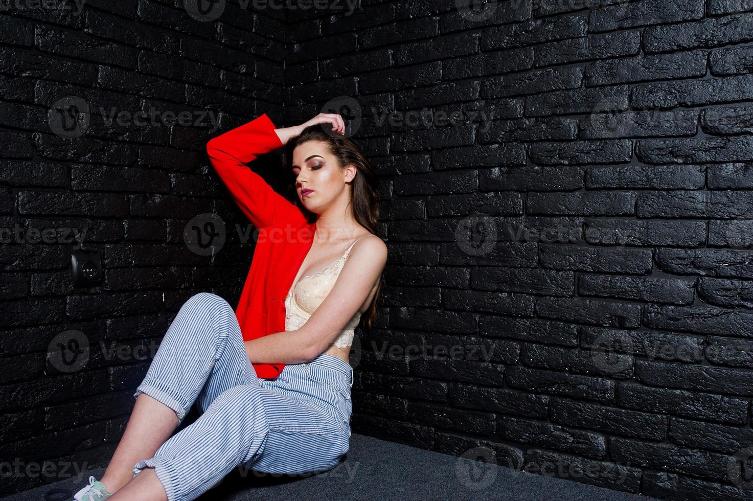 ragazza bruna alla moda sulla giacca rossa contro il muro di mattoni nero dello studio. foto