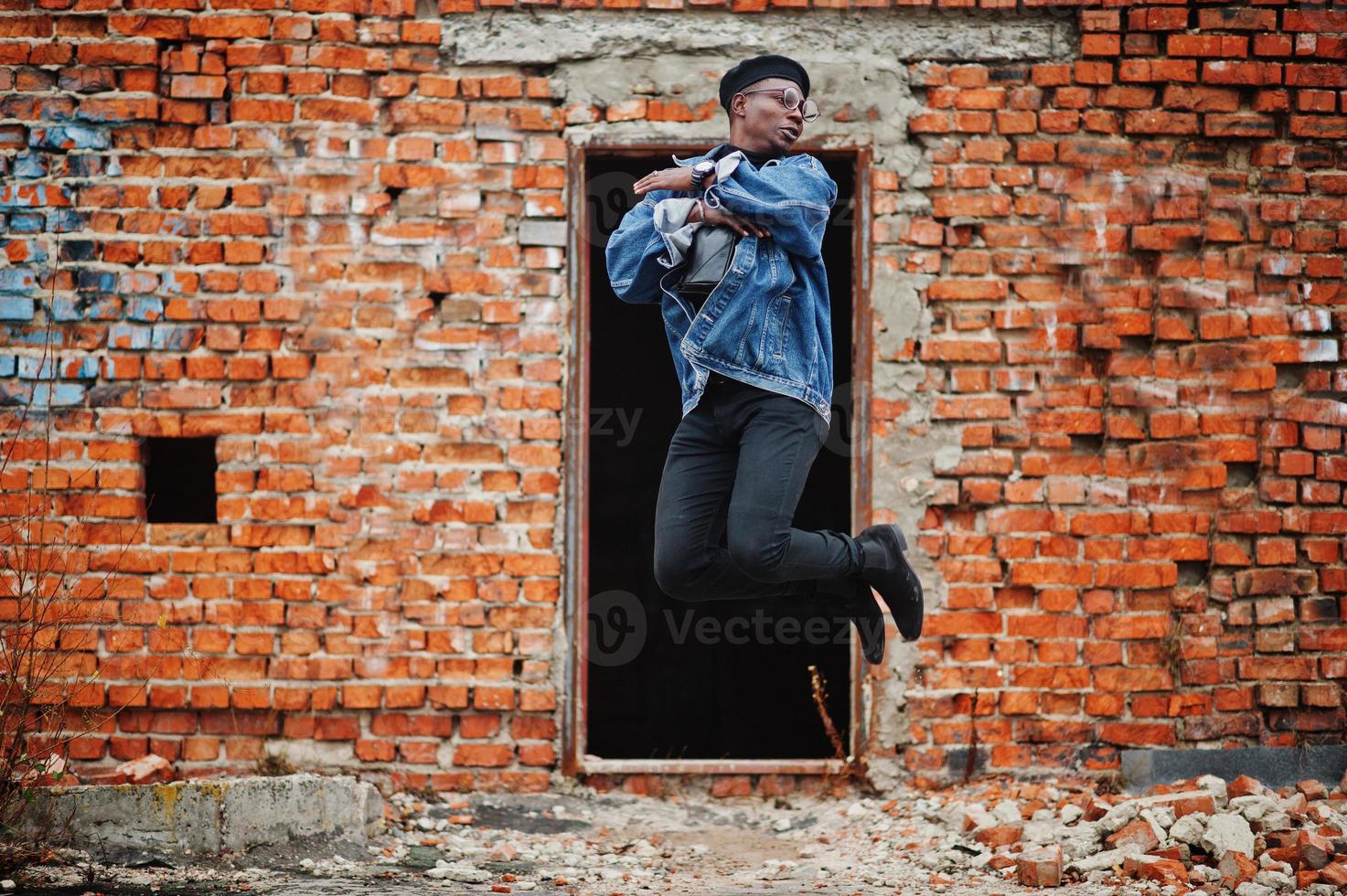 l'uomo afroamericano in giacca di jeans, berretto e occhiali da vista contro il muro di mattoni salta al tetto abbandonato. foto