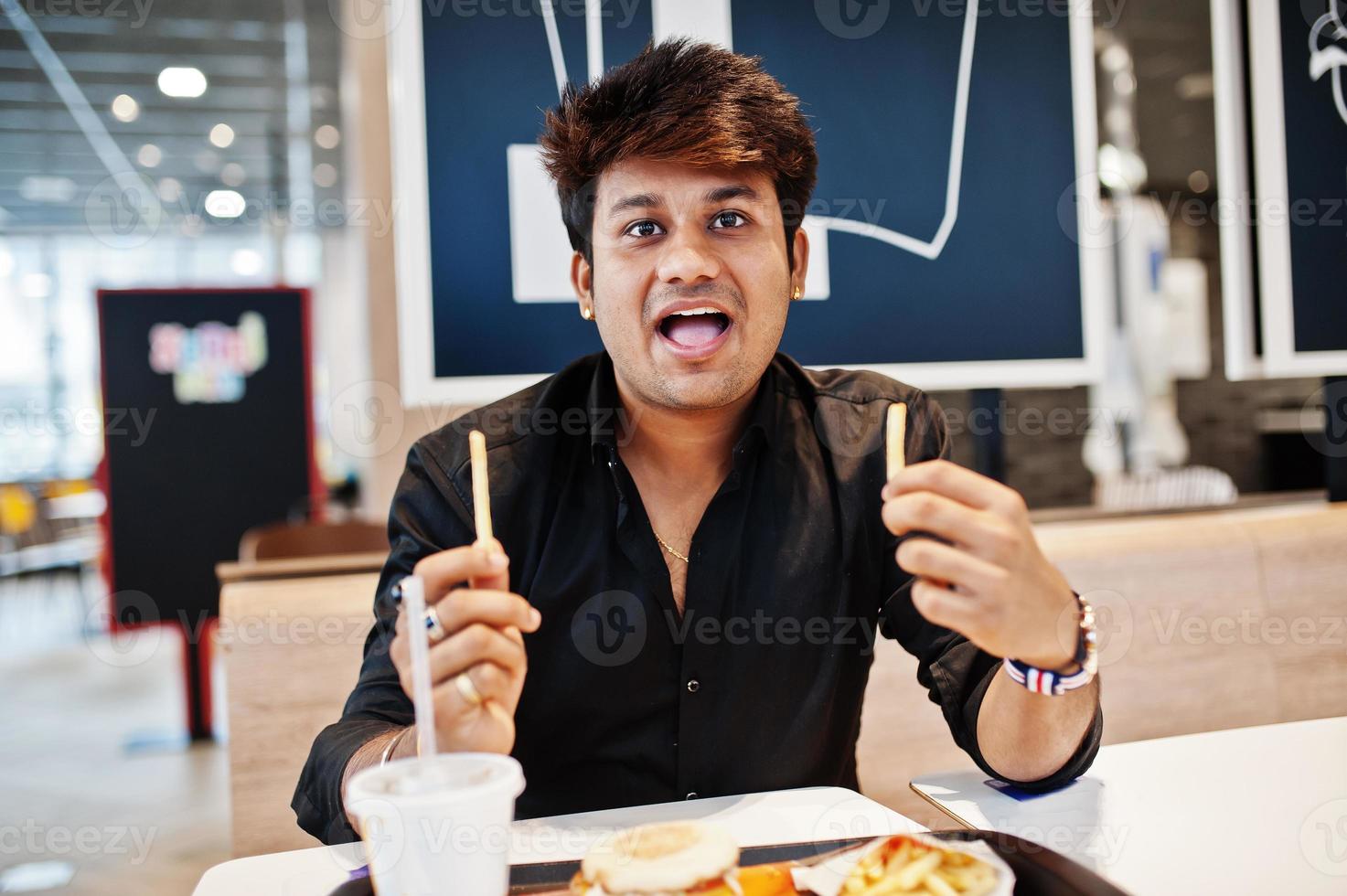uomo indiano divertente ed elegante seduto al fast food e mangiare patatine fritte, mostra la sua lingua. foto
