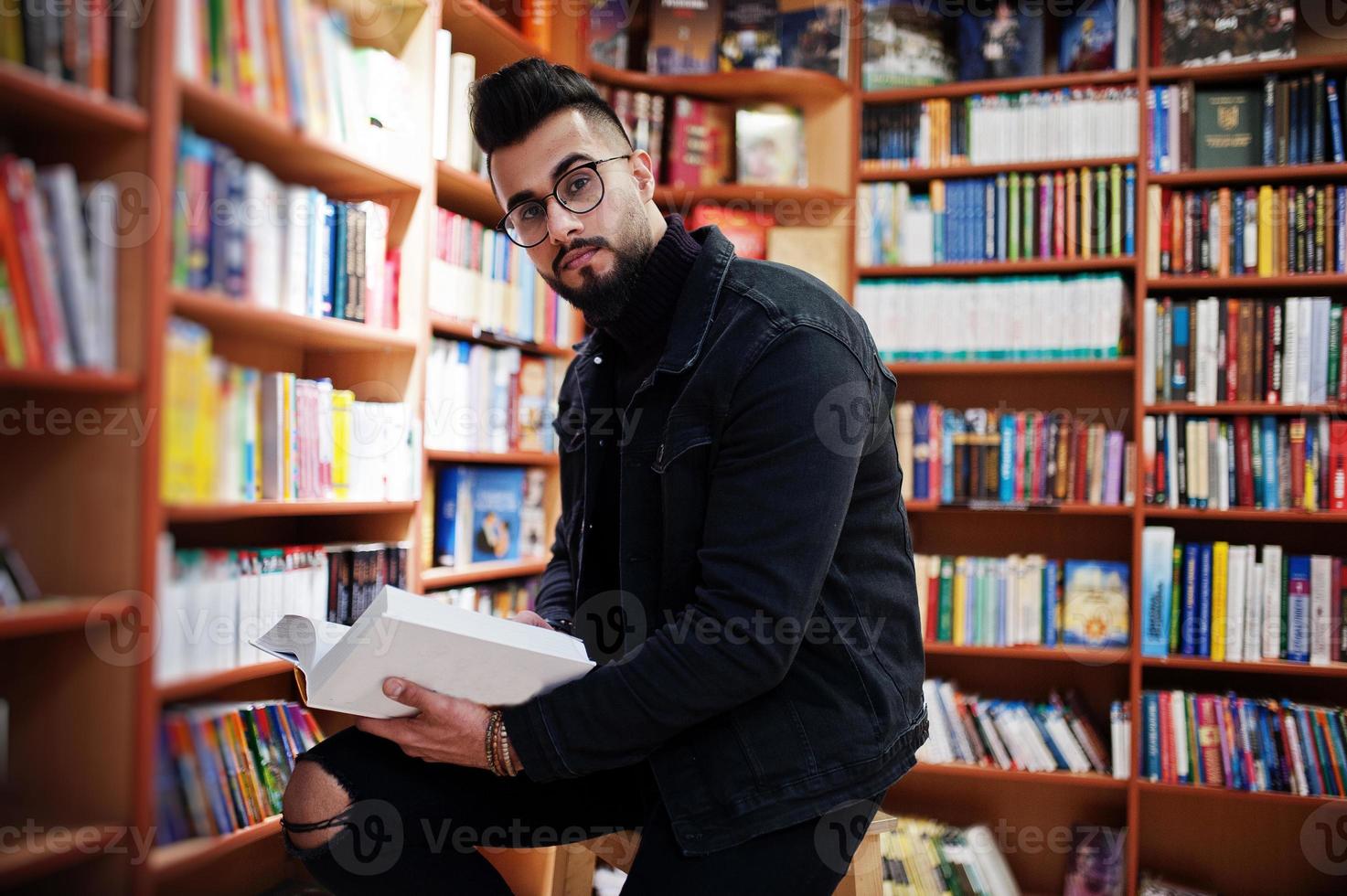 studente arabo alto e intelligente, indossa una giacca di jeans nera e occhiali da vista, in biblioteca con un libro a portata di mano. foto