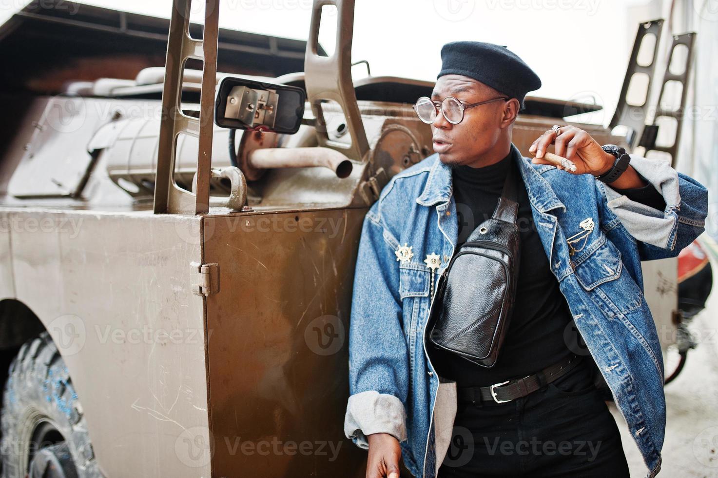 uomo afroamericano in giacca di jeans, berretto e occhiali, fumando sigaro e posato contro un veicolo blindato militare btr. foto