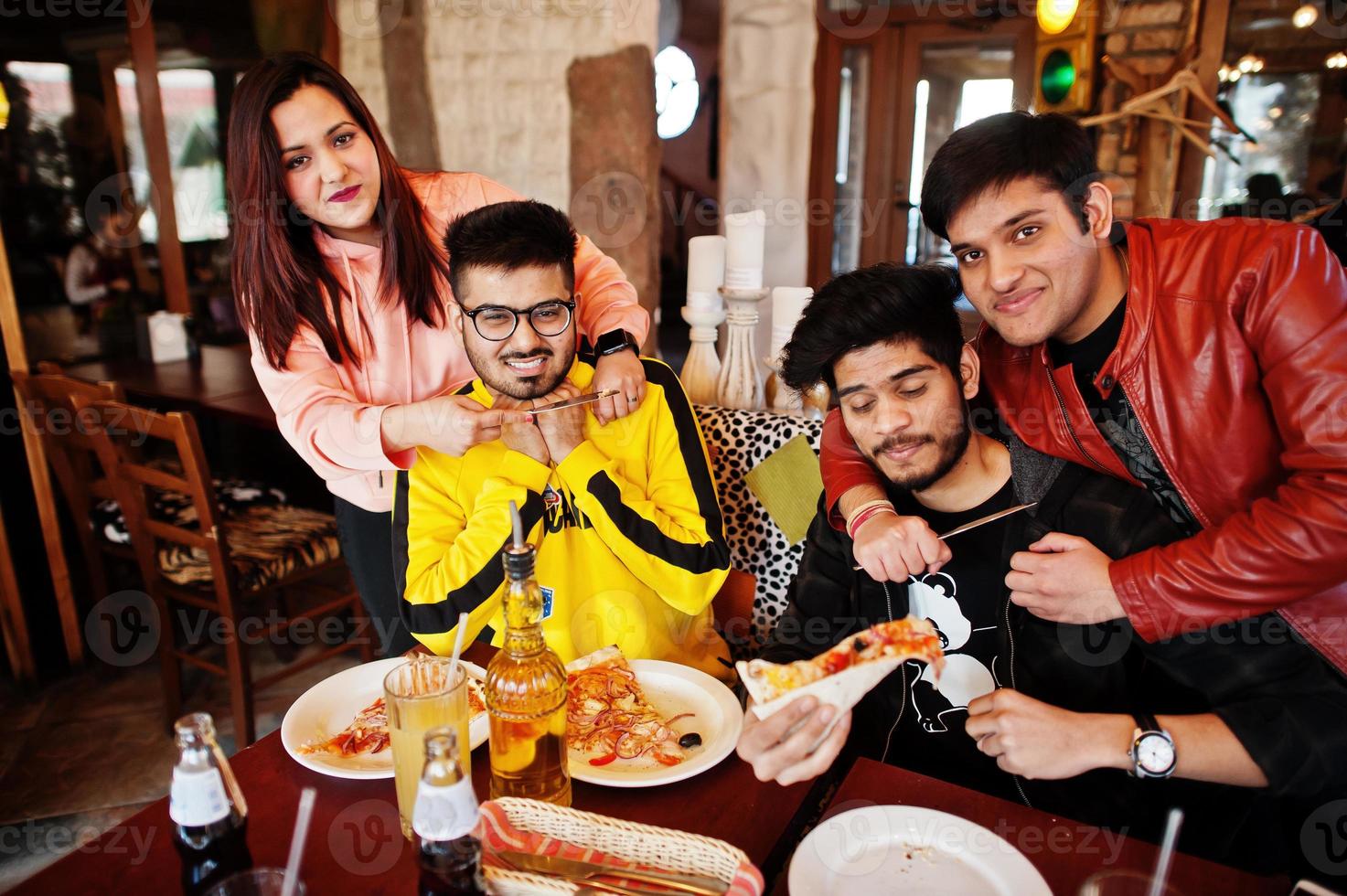 gruppo di amici asiatici che mangiano pizza durante la festa in pizzeria. felici indiani che si divertono insieme, mangiano cibo italiano e si siedono sul divano, litigando con forchette e coltelli. foto