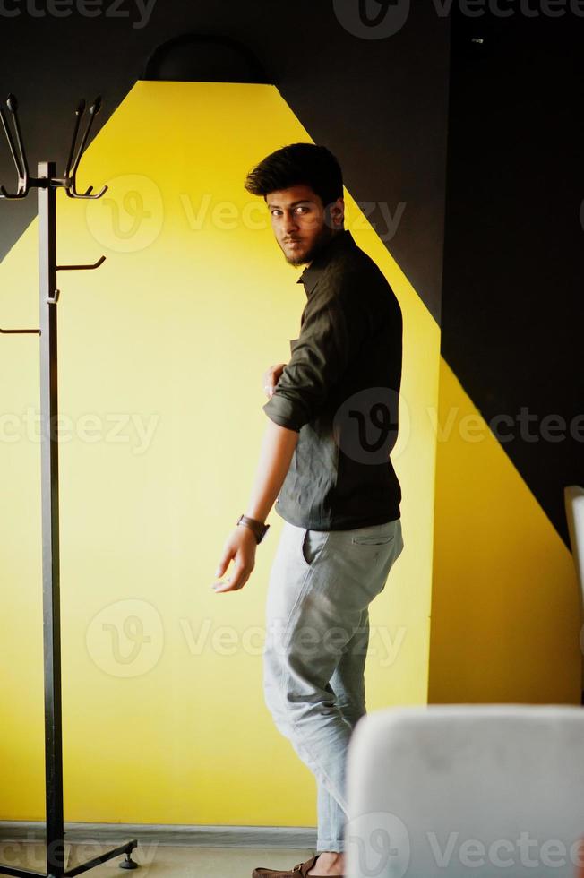 fiducioso giovane indiano in camicia nera in piedi vicino al muro giallo. foto