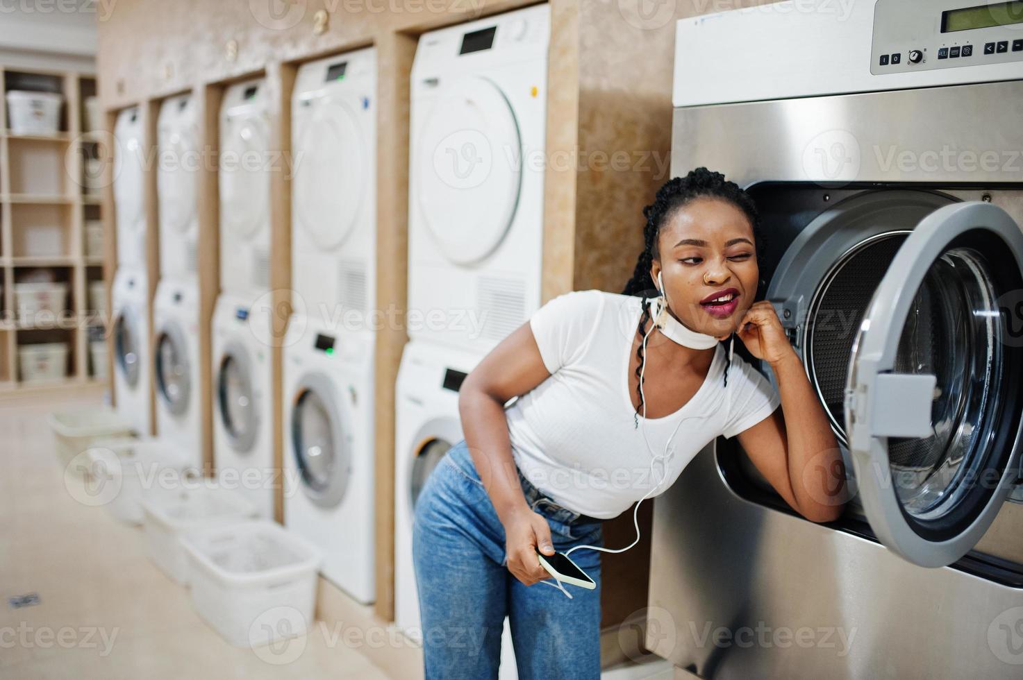allegra donna afroamericana vicino alla lavatrice ascoltando musica con gli auricolari dal telefono cellulare nella lavanderia self-service. foto