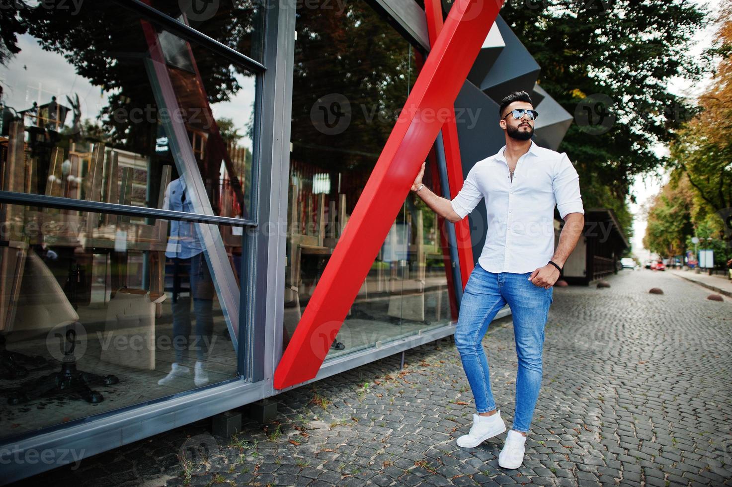 elegante uomo arabo alto modello in camicia bianca, jeans e occhiali da sole posato in una strada della città. barba attraente ragazzo arabo. foto