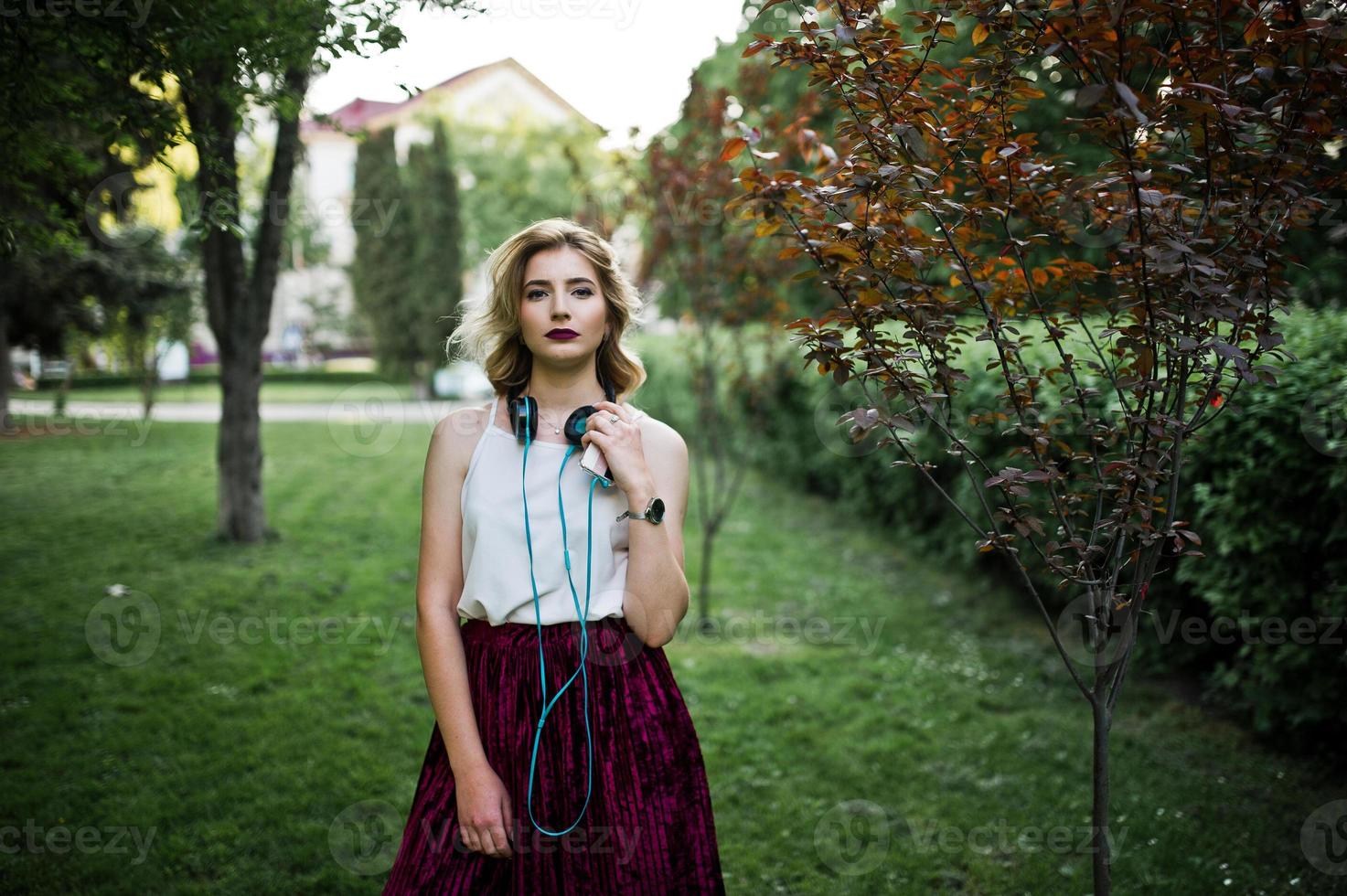 ragazza modello bionda alla moda e bella in elegante gonna di velluto di velluto rosso, camicetta bianca, posata al parco con telefono e auricolari. foto