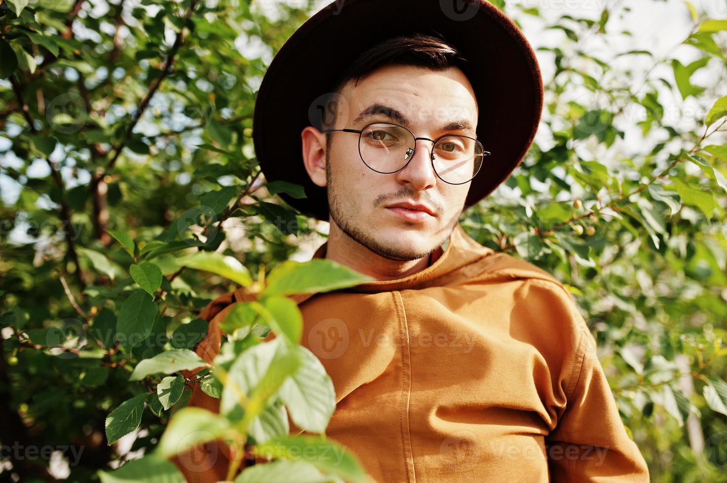 uomo alla moda con gli occhiali, giacca marrone e cappello in posa sul campo verde. foto