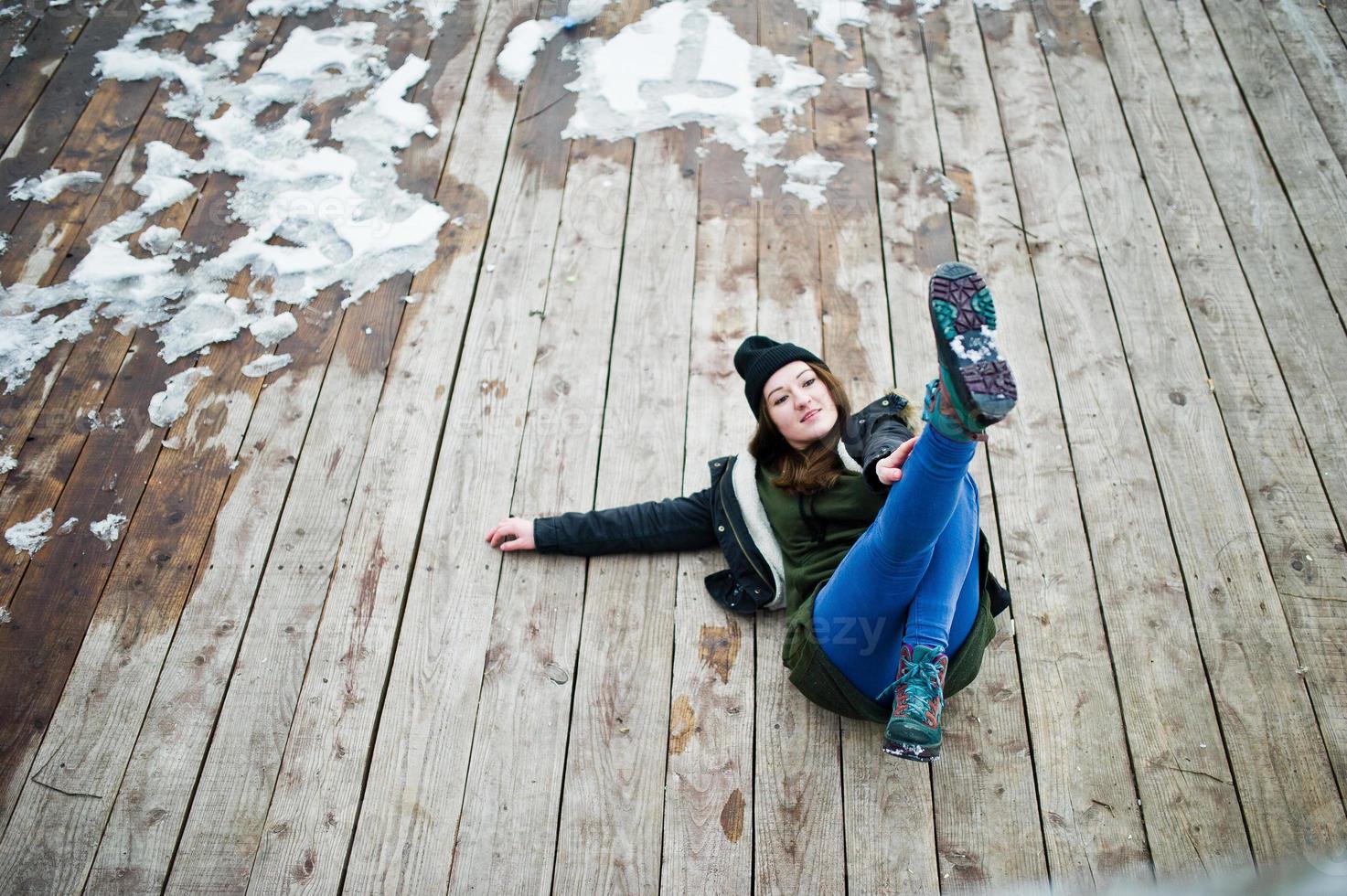 giovane ragazza indossa una lunga felpa verde, jeans e copricapo nero seduto sul pavimento di legno con la neve. foto