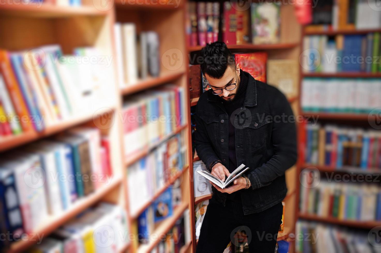 studente arabo alto e intelligente, indossa una giacca di jeans nera e occhiali da vista, in biblioteca con un libro a portata di mano. foto