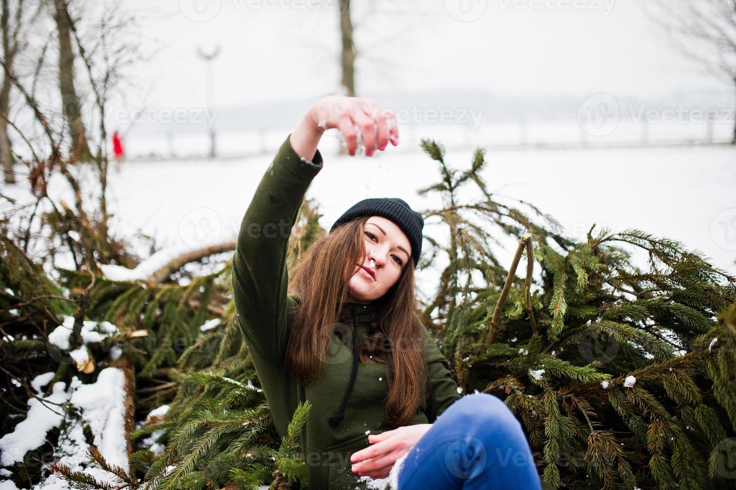 la ragazza indossa una lunga felpa verde, jeans e copricapo nero sui rami del pino nelle giornate invernali. foto