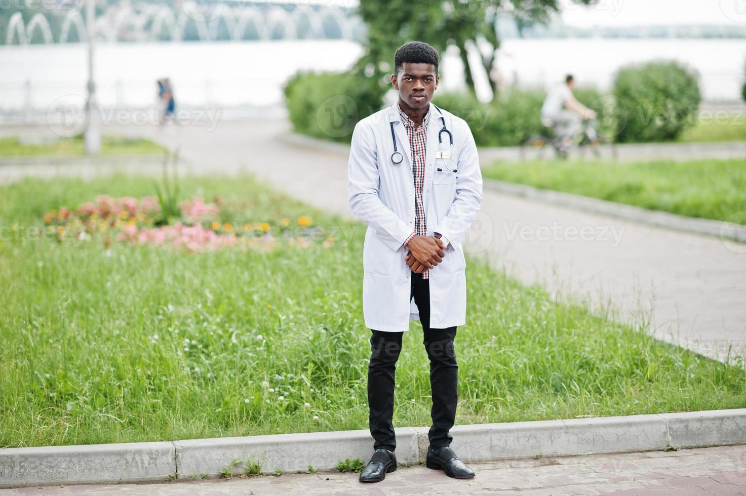 giovane medico maschio afroamericano in camice bianco con uno stetoscopio posato all'aperto. foto