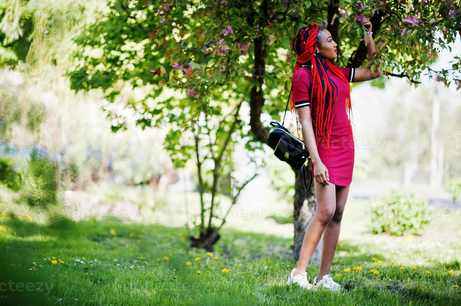 ragazza afroamericana carina e snella in abito rosso con dreadlocks poste all'aperto nel parco primaverile. modello nero elegante. foto