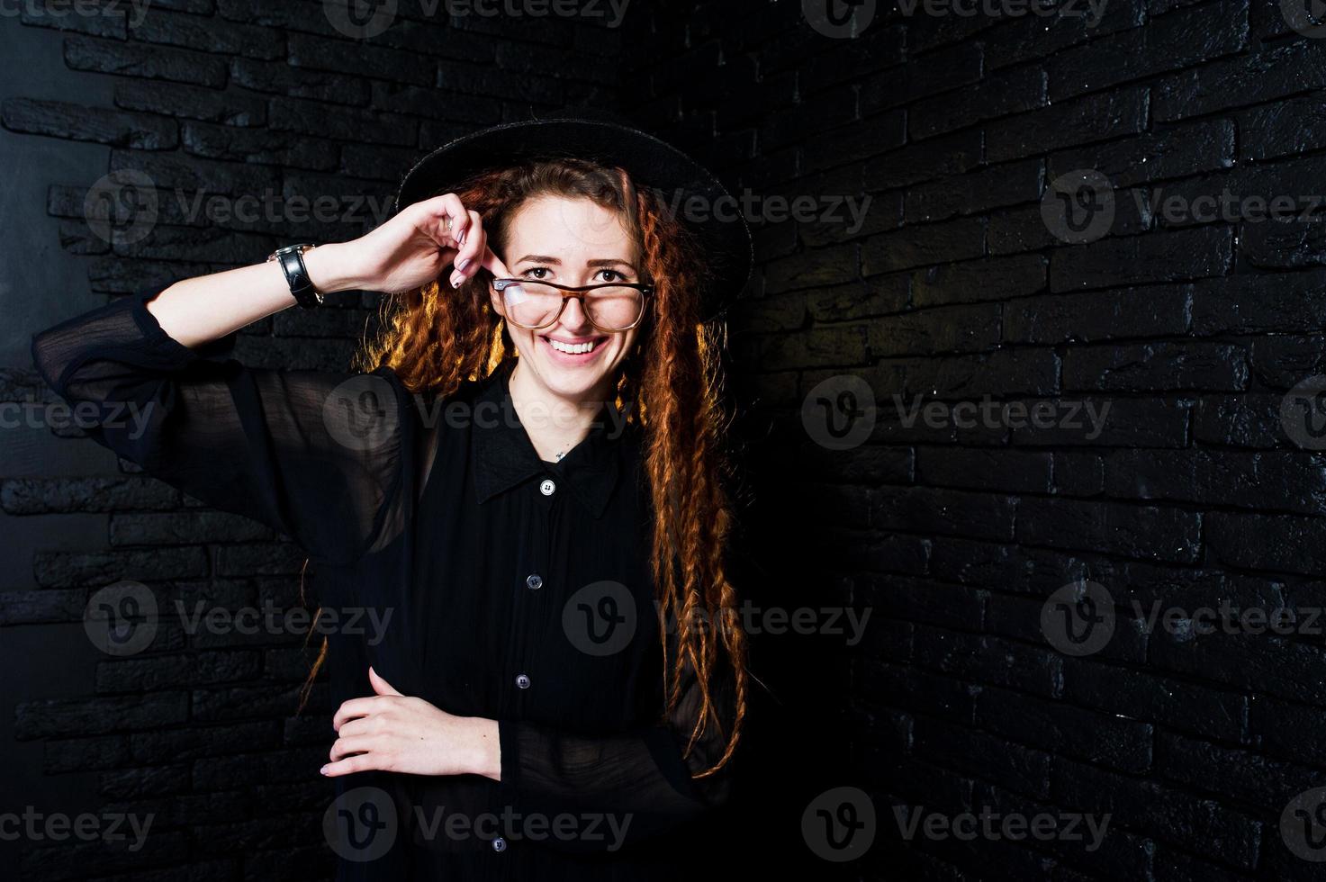 tiro in studio di una ragazza in nero con dreadlocks, occhiali e cappello su sfondo di mattoni. foto