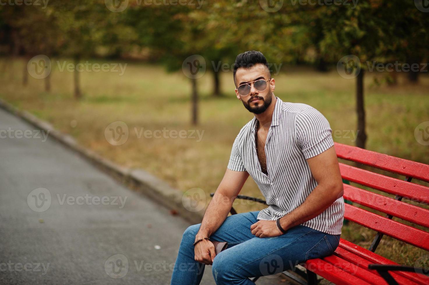l'uomo alla moda con barba araba alta indossa camicia, jeans e occhiali da sole seduto su una panchina rossa al parco. foto