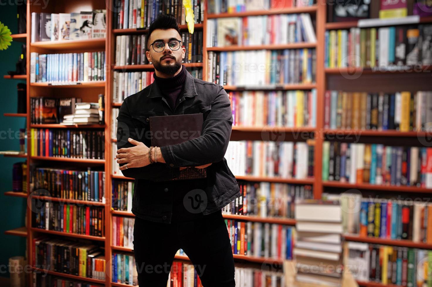 studente arabo alto e intelligente, indossa una giacca di jeans nera e occhiali da vista, in biblioteca con un libro a portata di mano. foto