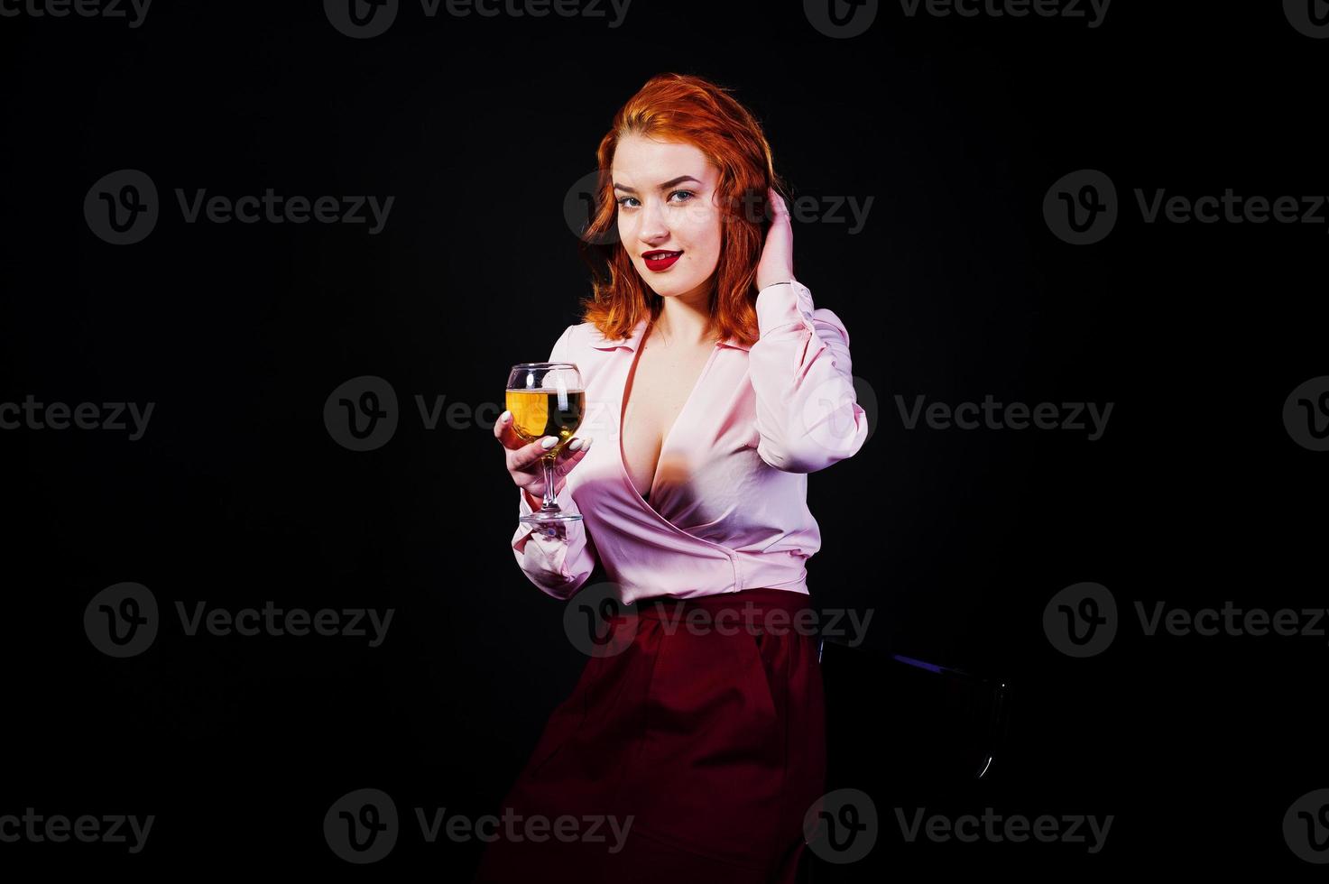splendida ragazza dai capelli rossi in camicetta rosa e gonna rossa con un bicchiere di vino a portata di mano isolato su nero. foto