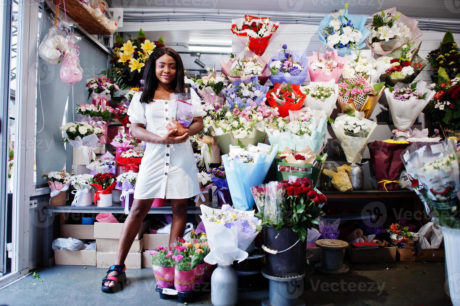 bella ragazza afroamericana in tenero abito bianco con bouquet di fiori in mano in piedi su sfondo floreale nel negozio di fiori fiorista femminile nero. foto