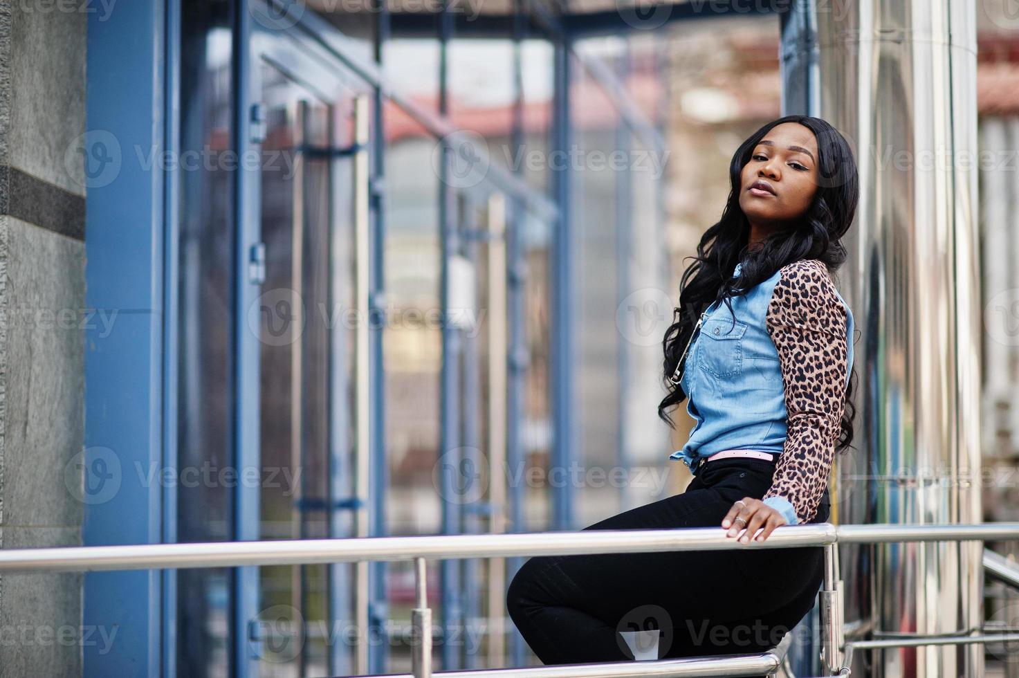 hipster ragazza afroamericana che indossa una camicia di jeans con maniche di leopardo in posa in strada contro un moderno edificio per uffici con finestre blu. foto