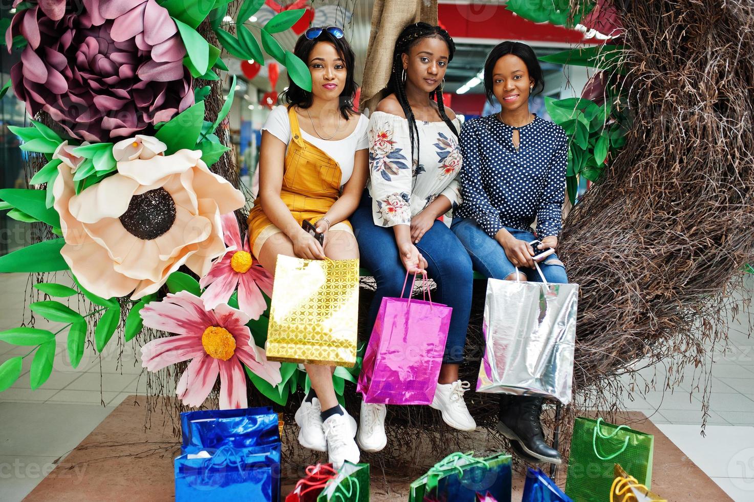 belle tre ragazze afro americane ben vestite con borse della spesa colorate sedute su una zona fotografica di decorazione primaverile nel centro commerciale. foto