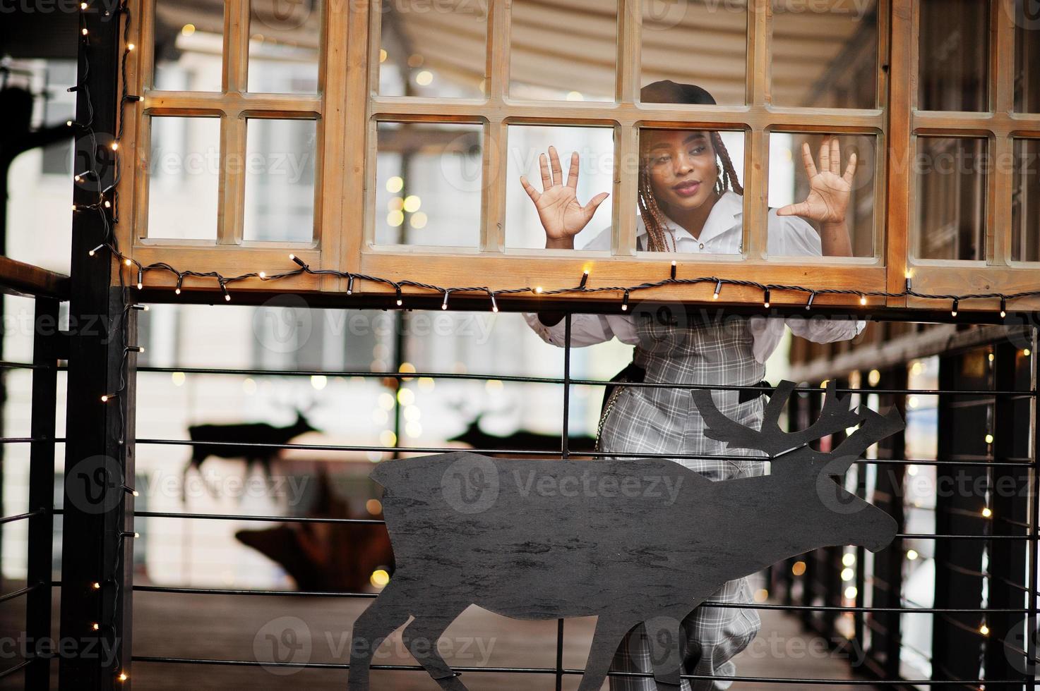 donna afroamericana in tuta e berretto poste in terrazza all'aperto con decorazioni natalizie ghirlanda e cervi. foto