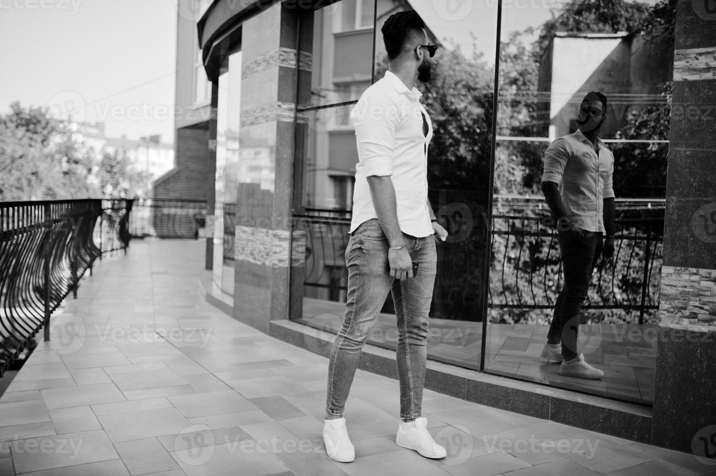 elegante uomo arabo alto modello in camicia bianca, jeans e occhiali da sole posato in una strada della città. barba attraente ragazzo arabo contro edificio moderno. foto