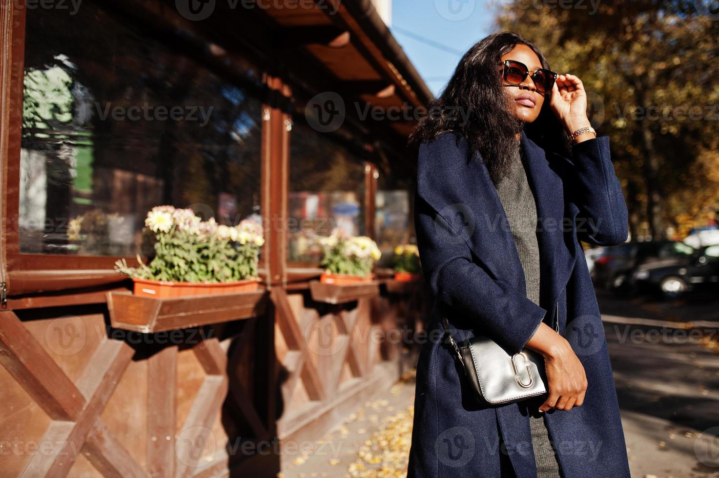 elegante ragazza afroamericana in cappotto blu e occhiali da sole poste in una soleggiata giornata autunnale. donna modello africano. foto