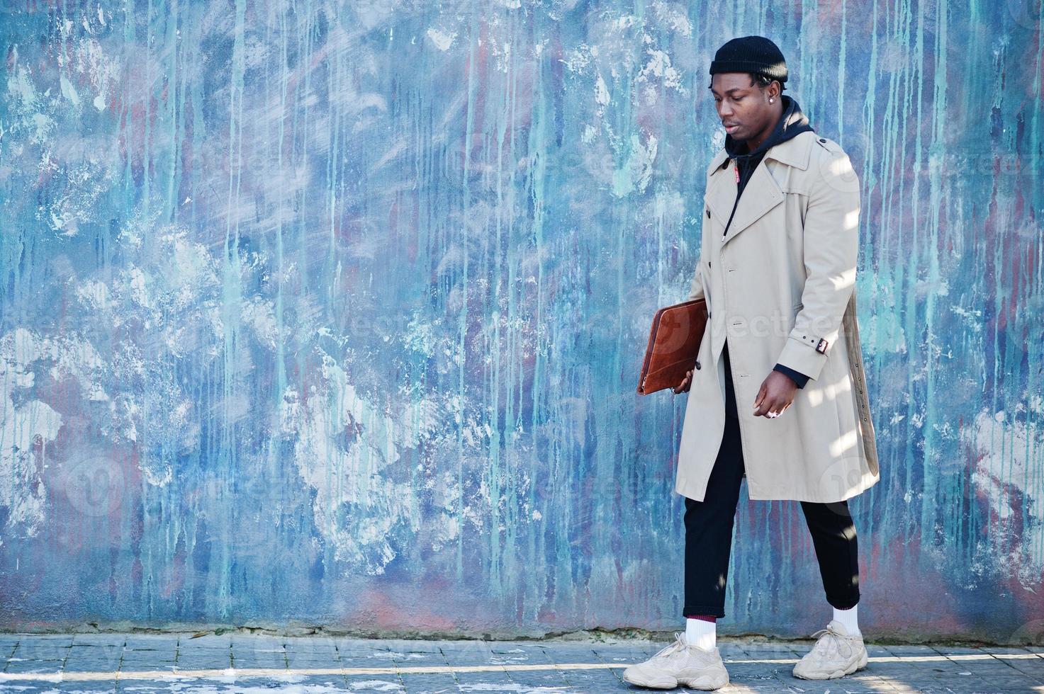 bell'uomo afroamericano in posa fuori con cappello nero e cappotto beige con cartella in mano. foto