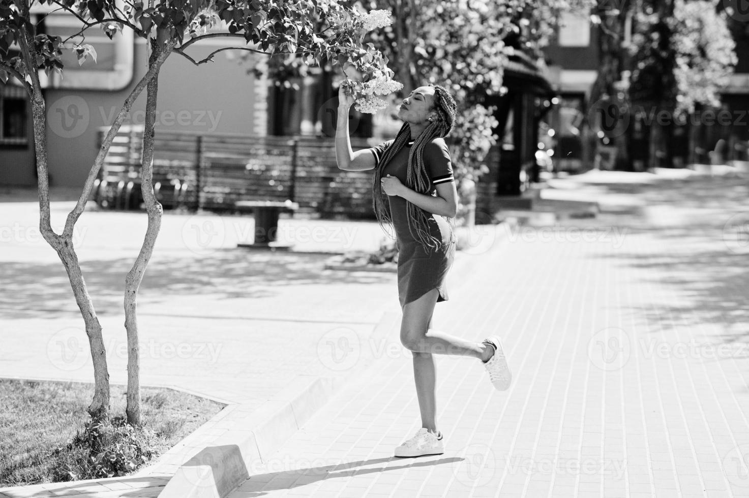 ragazza afroamericana carina e snella in abito rosso con dreadlocks poste all'aperto sulla strada vicino all'albero lilla. modello nero elegante. foto