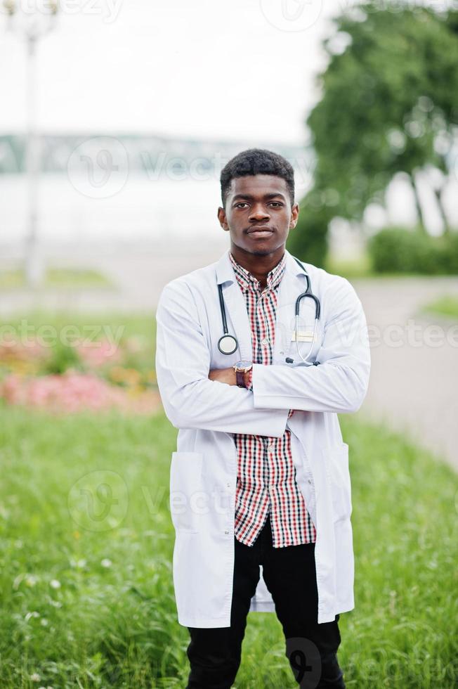 giovane medico maschio afroamericano in camice bianco con uno stetoscopio posato all'aperto. foto